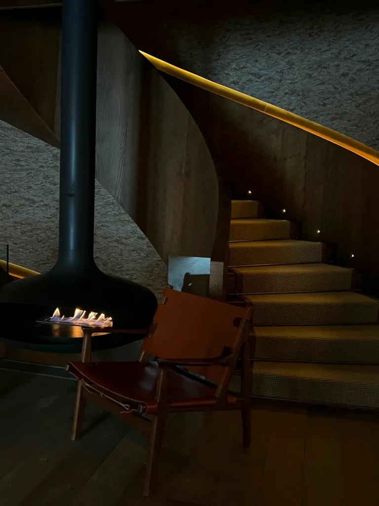 Chimenea suspendida encendida, una butaca de cuero y una escalera curva en penumbra