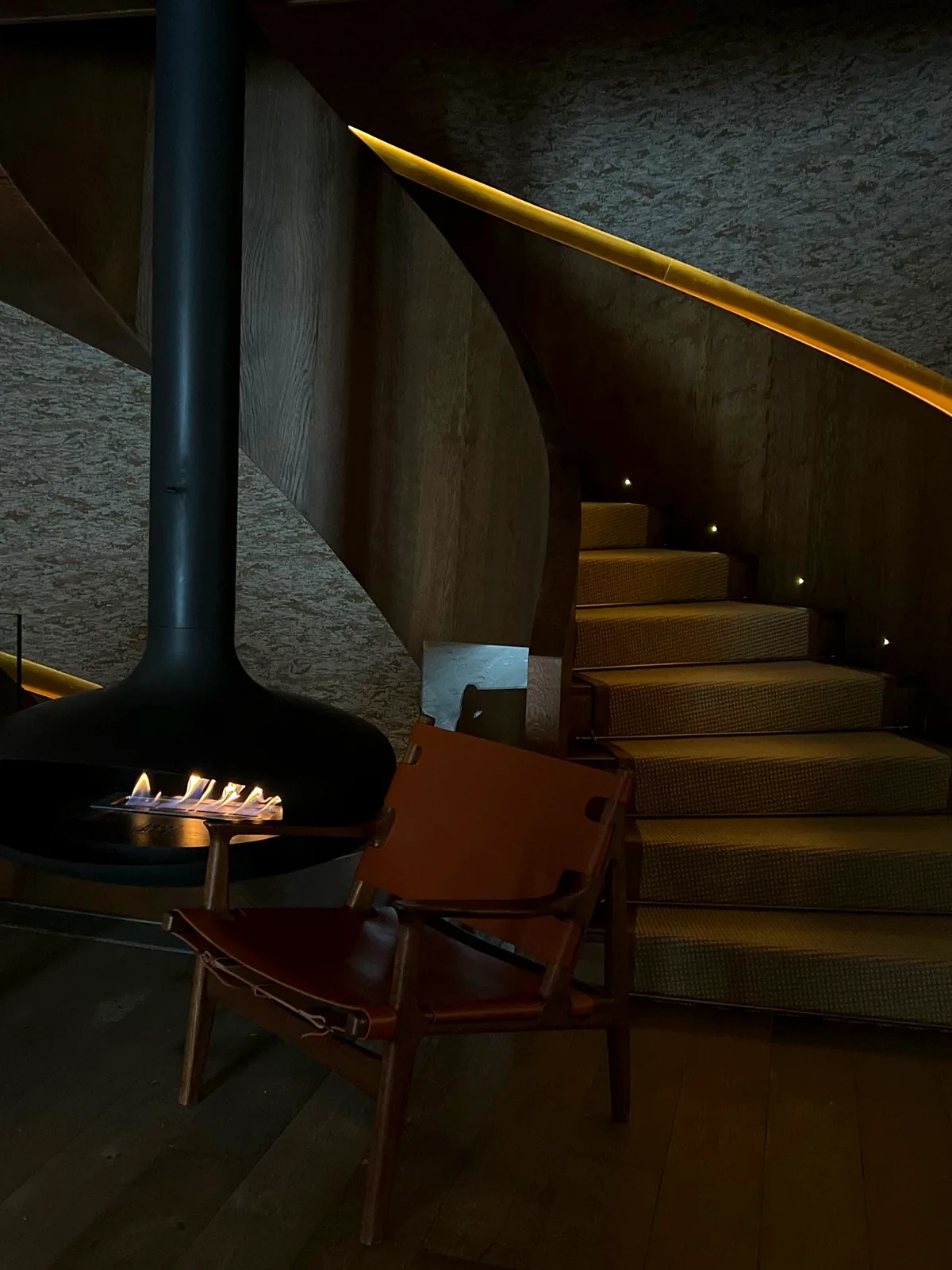 Chimenea suspendida encendida, una butaca de cuero y una escalera curva en penumbra