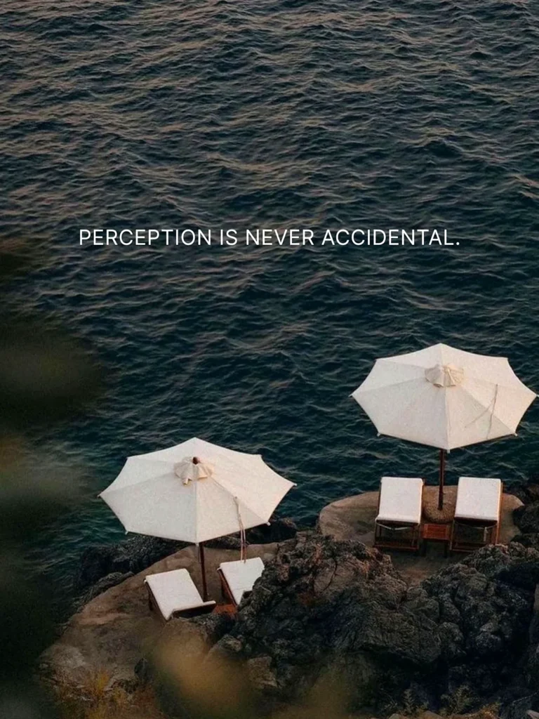 Sombrillas blancas y tumbonas sobre unas rocas junto al mar, con el texto «PERCEPTION IS NEVER ACCIDENTAL.»